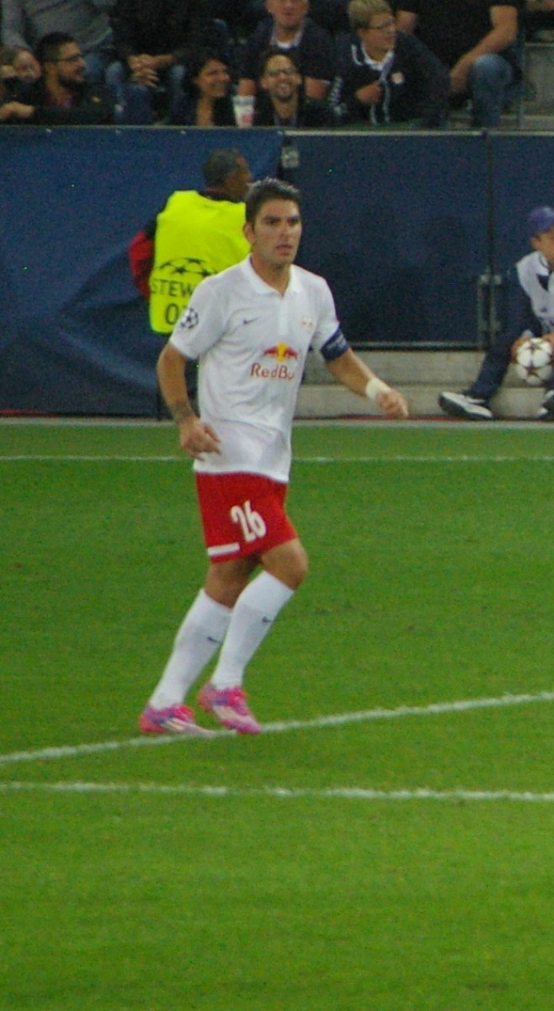 Ein professioneller Fußballspieler in weißem Trikot und roter Hose kickt einen Ball auf einem Feld, mit Zuschauern im Hintergrund.