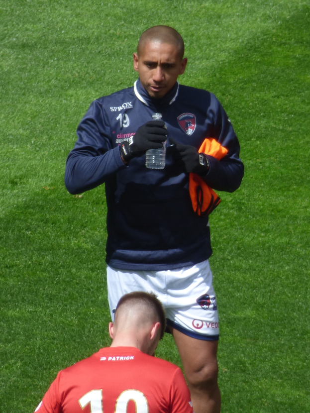 Ein professioneller Sportler in einem Sportoutfit steht auf einem saftigen grünen Feld, in einer Hand eine Flasche und in der anderen ein oranges Tuch haltend.