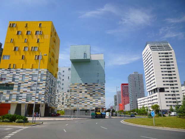 Eine Stadtstraße mit hohen Gebäuden im Hintergrund, Straßenlaternen, Autos, Infotafeln, Büschen, Bäumen und einem bewölkten Himmel, mit einzigartig gestalteten Gebäuden der Universität Rotterdam.