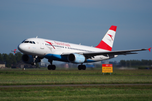 Airbus A320-200 der Austrian Airlines beim Abheben vom Frankfurt Airport, mit einem Schild im Vordergrund, Gras, Bäumen, Gebäuden und einem klaren blauen Himmel im Hintergrund.