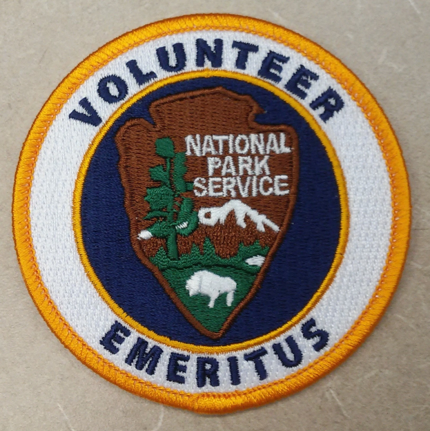 Rundes Abzeichen mit blauem Rand und weißem Hintergrund, darauf in schwarzer Schrift 'Volunteer National Park Service Emeritus' in der Mitte eines gelben Kreises.