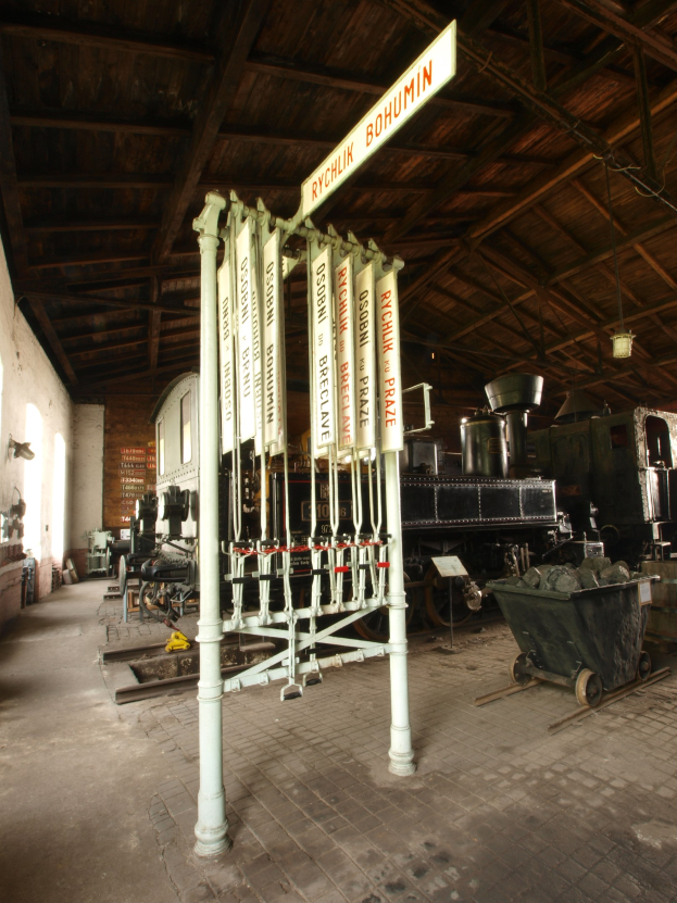 Ein Bahnsteig mit einem Zug auf den Schienen, einem Rollstuhl auf dem Boden, einer an einem Pfahl befestigten Tafel, einer Wand auf der linken Seite und einer Decke darüber sowie einer zentralen Signalbox zur Steuerung der Zugankünfte.