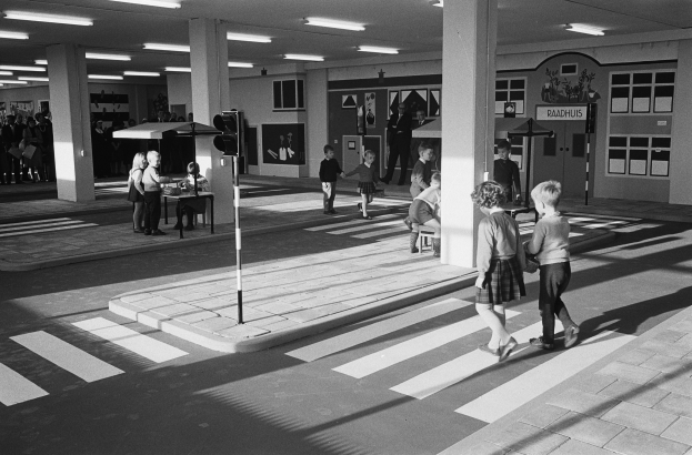 Schwarzes und weißes Foto von Kindern, die eine Straße an einer Ampel überqueren, mit Fußgängern in der Nähe, einem Verkehrssignalmast im Vordergrund und Architekturpfeilern mit Deckenleuchten im Hintergrund.
