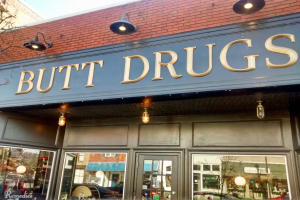 Außenansicht eines Gebäudes mit beleuchteter Schilder "butt drugs", das Glaswände und -türen hat, mit Fahrzeugen, Gebäuden, Bäumen und Himmel im Hintergrund.