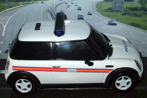 Weißes Polizeiauto vor einer belebten Straße mit mehreren Fahrzeugen, Gras und Bäumen auf beiden Seiten, aufgenommen außerhalb der Stadt.