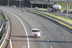 Ein Auto fährt auf einer Autobahn mit einem Polizeiwagen am Rande, anderen Fahrzeugen, Geländern, Polen, Planken, Schildern und Bäumen im Hintergrund.