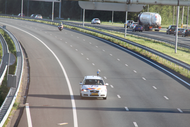 Ein Auto fährt auf einer Autobahn mit einem Polizeiwagen am Rande, anderen Fahrzeugen, Geländern, Polen, Planken, Schildern und Bäumen im Hintergrund.
