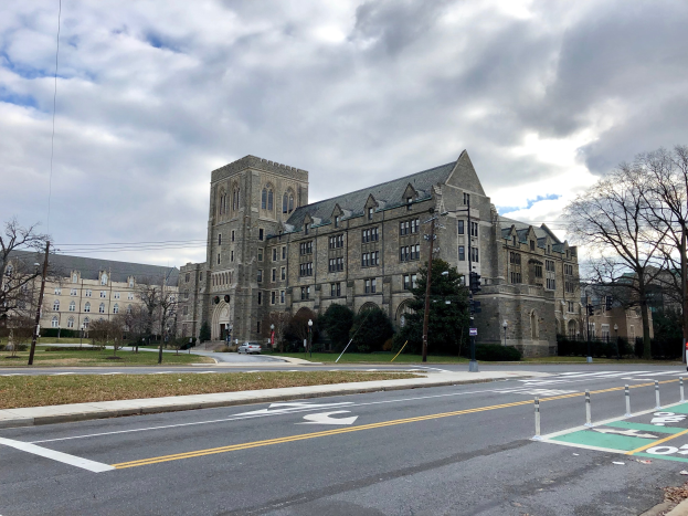 Großes Gebäude mit vielen Fenstern, als Universität von Pennsylvania beschriftet, umgeben von Bäumen, Straßenlaternen, Verkehrszeichen und Fahrzeugen an einem bewölkten Tag.