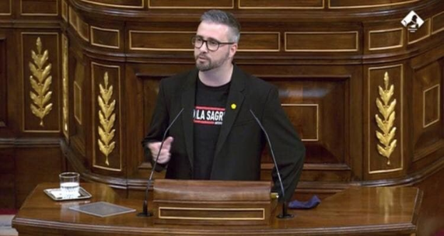 Ein Mann in einem schwarzen Blazer und Brille steht an einem Rednerpult mit zwei Mikrofonen, einem Glas Wasser, einem Buch und einem blauen Tuch und hält eine Rede als Führer der sozialistischen Partei Frankreichs.