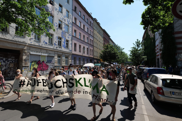 Eine Gruppe von Menschen, die mit einem Banner auf der Straße marschieren, mit parkenden Autos auf der rechten Seite und einem Radfahrer auf der linken Seite, vor Gebäuden und Bäumen während einer Demonstration in Berlin.