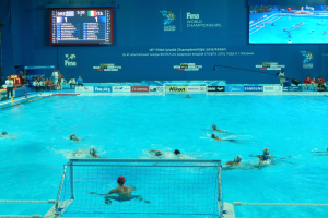 Eine Gruppe von Menschen spielt Wasserball in einem großen Pool mit einem Netz in der Mitte, Zuschauern auf der linken Seite und einem Bildschirm mit Text und Logos im Hintergrund bei den FINA-Weltmeisterschaften 2016 in Kazan.