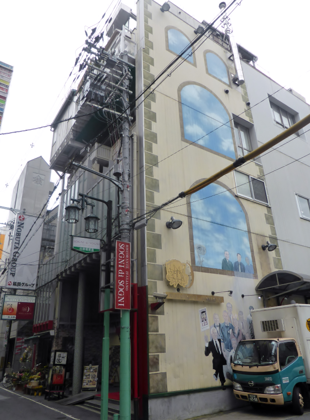 Ein Truck steht auf der Straßenseite neben einem hohen Gebäude mit einer Wandmalerei, mit Strommasten, Kabeln, Straßenlaternen, Namensschildern, Kraftfahrzeugen, Topfpflanzen und einem klaren blauen Himmel im Hintergrund.