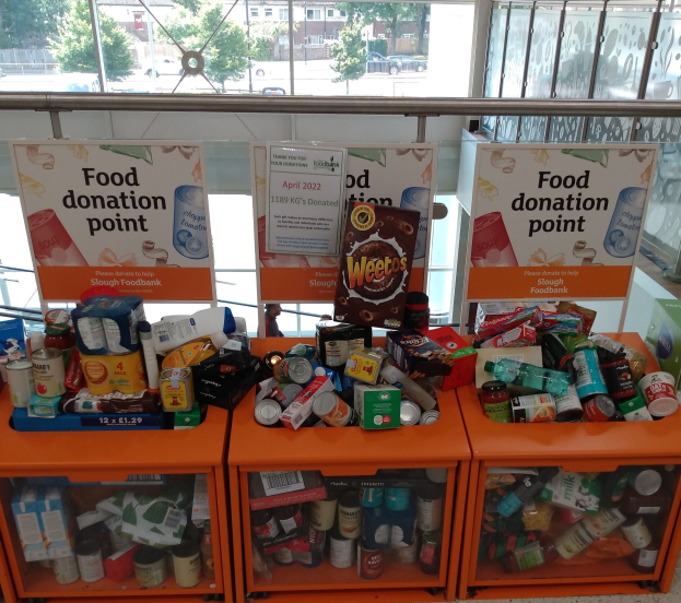 Eine Spendenstation mit orangefarbenen Schränken, die Flaschen, Dosen, Kartons und andere Gegenstände enthalten, begleitet von Texttafeln, betrachtet durch Glasfenster, die Fahrzeuge, Bäume, Gebäude und Himmel zeigen.