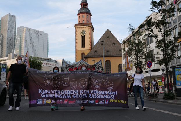 Eine Gruppe von Menschen mit Masken, die eine Straße entlang marschieren und ein Banner halten, mit einem geparkten Auto auf der linken Seite, Gebäuden, Bäumen, Schildern, Polen, einem Kirchturm und einem klaren blauen Himmel im Hintergrund.