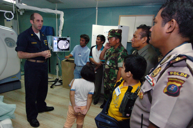 Eine Gruppe von Menschen versammelt sich um ein Krankenhauszimmer mit einem Mann, der ein Mikrofon hält, einem drapierten Bett auf der linken Seite, medizinischen Geräten im Hintergrund, einem Röntgenmaschinenständer, einer Texttafel, einer Tür und Deckenleuchten.