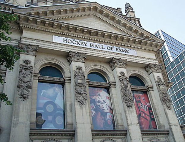 Außenansicht des Hockey Hall of Fame in New York City, mit Glasfenstern, Text auf dem Gebäude, einem Baum links und einem sichtbaren Himmel.