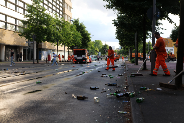 Menschen in orangefarbenen Uniformen sammeln Müll von einer Straße mit Flaschen und Schutt, mit Bäumen, Pfählen und Fahrzeugen im Hintergrund.