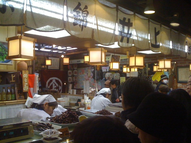 Restaurantmitarbeiter bereiten Artikel hinter einer Theke vor, während Kunden davor stehen, mit Sauceflaschen, Menüs, Kleidung und Lampen im Hintergrund.