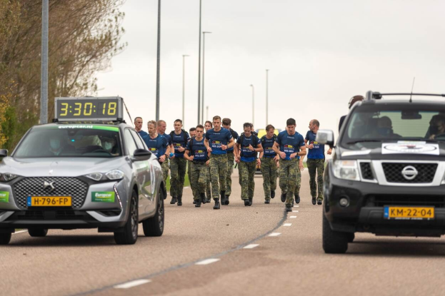 Gruppe von Menschen, die auf einer Straße neben geparkten Autos gehen, mit Bäumen auf der linken Seite und Strommasten im Hintergrund, unter einem sichtbaren Himmel.