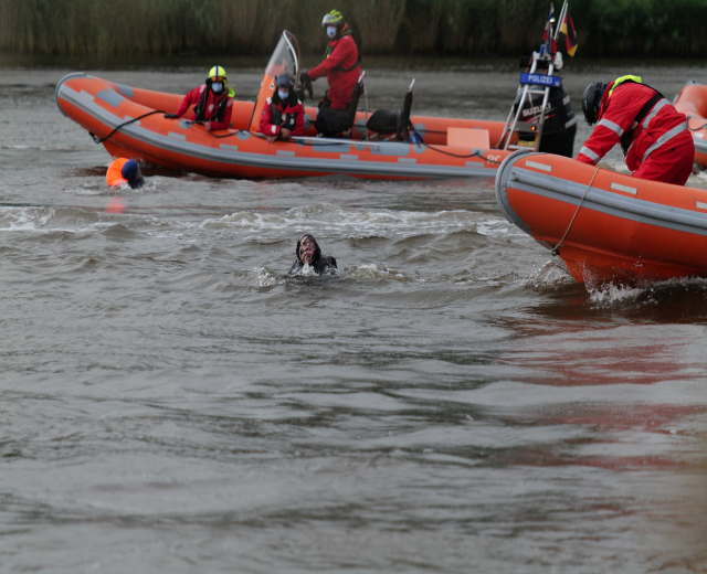 Gruppe von Menschen in einem aufblasbaren Boot auf einem Fluss, mit zwei Personen im Wasser im Vordergrund und Vegetation im Hintergrund, alle tragen Schwimmwesten und Helme während einer Rettungsoperation.