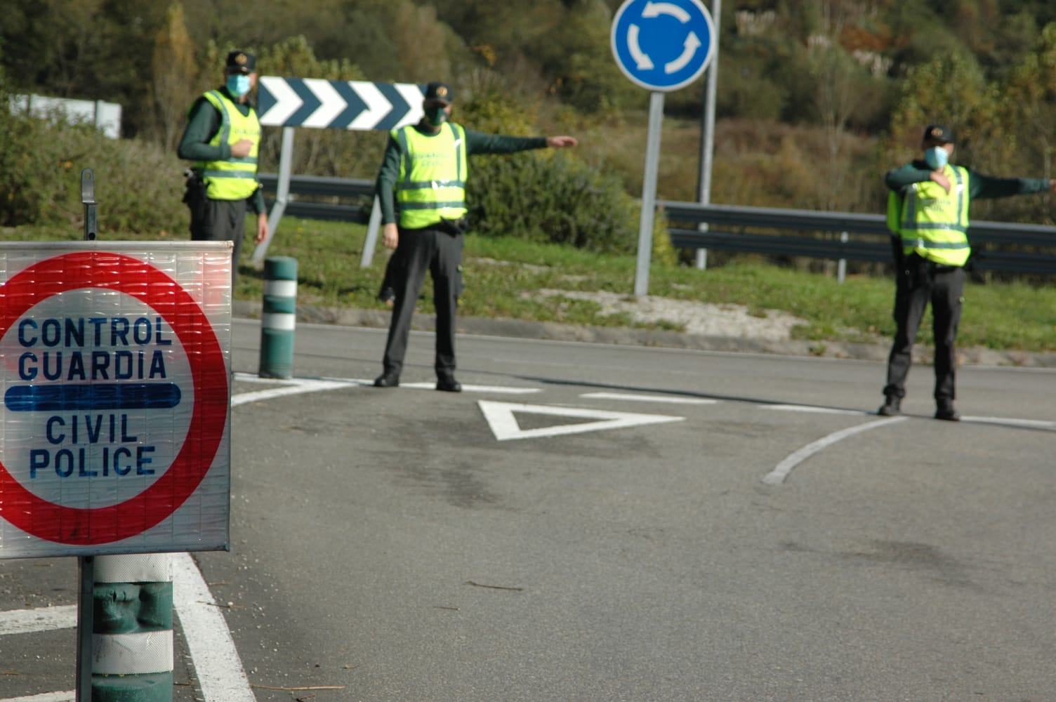 Drei Polizisten in grünen Jacken, schwarzen Hosen und schwarzen Schuhen stehen an der Seite einer Straße mit einem Verkehrsschild und einem Laufschuh im Vordergrund und Bäumen, Pflanzen, Gras und einem Geländer im Hintergrund.