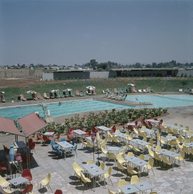 Ein Freibad mit Tischen, Stühlen und Sonnenschirmen umgeben von Pflanzen, einem Zaun, einer Wand, Bäumen und Gebäuden unter einem bewölkten Himmel.