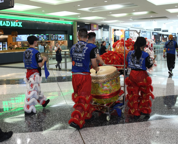 Eine Gruppe von Menschen in roter und blauer Kleidung, die durch einen Flughafen gehen, einige mit Musikinstrumenten und Gep├Ącken, mit Gesch├Ąften und beleuchteter Decke im Hintergrund.