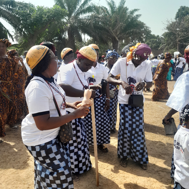 Eine Gruppe von Frauen in weißen und schwarzen Kleidern, einige mit gelben Helmen, steht zusammen auf einem staubigen Feld mit Bäumen und einem klaren blauen Himmel im Hintergrund, was auf ein Gemeindeveranstaltungs-Event in Ghana hinweist.