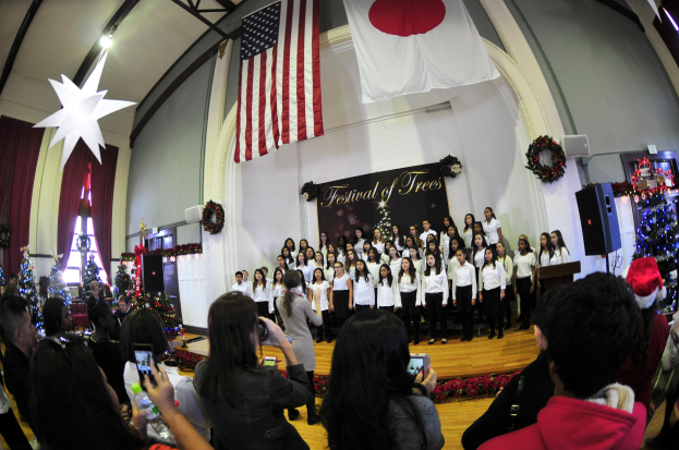 Eine Gruppe von Menschen auf einer Bühne mit Weihnachtsdekoration, einem Podium, Lautsprechern, einer Texttafel und einem Publikum.