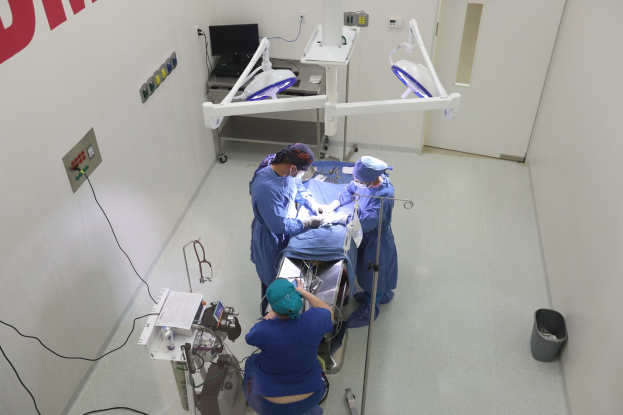 Eine Gruppe von Chirurgen führt einen Eingriff in einem Operationssaal durch, wobei ein Patient auf einem Operationstisch umgeben von medizinischen Geräten liegt, eine textbedeckte Wand auf der linken Seite und verschiedene medizinische Geräte im Hintergrund.