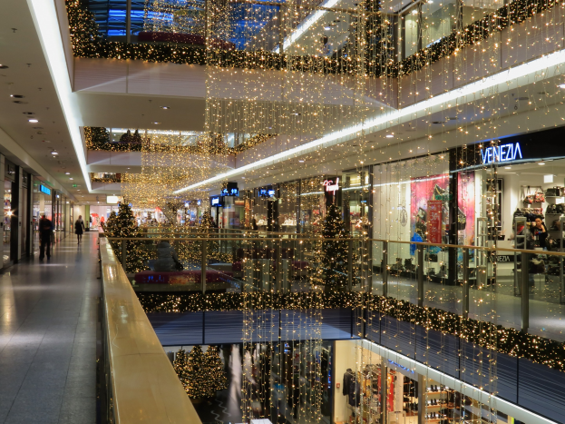 Das Innere eines festlich geschmückten Einkaufszentrums mit Weihnachtsbeleuchtung, das Menschen zeigt, die herumlaufen, Geschäfte mit Schildern, Glasgeländer und von der Decke hängende Deko, die eine einladende Weihnachtsatmosphäre schafft.