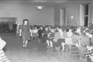 Eine Frau in einem schwarzen und weißen Kleid läuft über einen Laufsteg während einer Modenschau für Frauenwahlrecht, mit einer Menge, die sitzt und zusieht, einer Uhr und Lampen an der Wand und einer beleuchteten Decke.