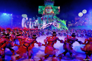 Eine Gruppe von Menschen in bunten Kostümen tanzt auf einer Bühne vor einem sitzenden Publikum, mit Lichtern, Polen, Ballons und einem Bildschirm, der eine Person in den Hintergrund zeigt, unter einem sichtbaren Himmel, bei der Rio-Karneval 2014.