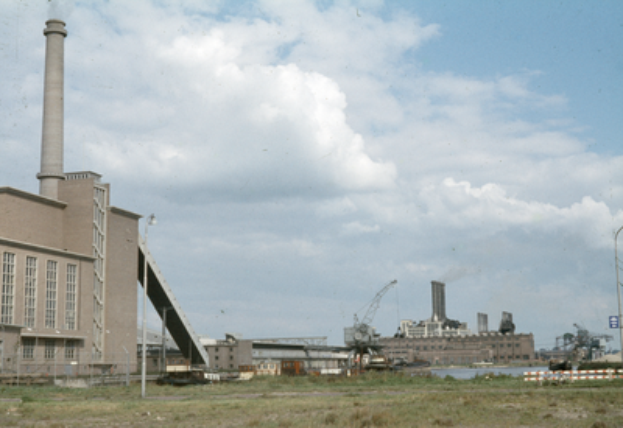 Ein altes Foto einer Fabrik mit Rauch, der aus ihren Schornsteinen quillt, umgeben von verschiedenen Industriegebäuden, mit grasbewachsenem Boden und einem bewölkten Himmel.
