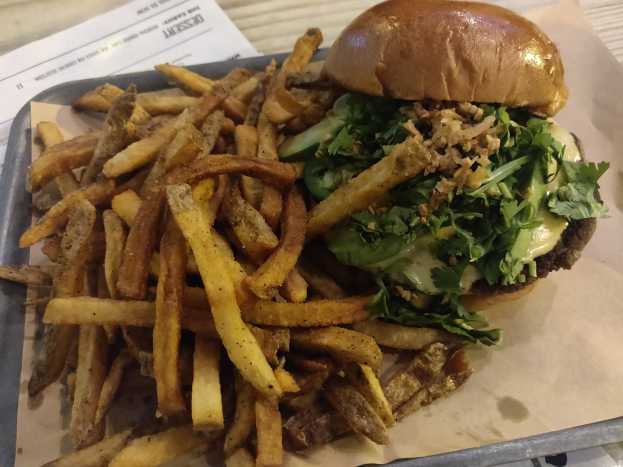 Ein Tablett mit einem Hamburger, der mit Salat, Tomate und Zwiebeln garniert ist und goldenen, knusprigen Pommes frites, das auf einem Tisch mit einem Blatt daneben steht.