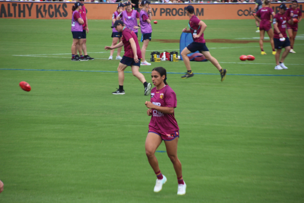 Frauen beim Australian Rules Football auf einem grünen Feld mit verstreuten Bällen und einem Banner im Hintergrund, einige tragen Kappen und Turnschuhe.