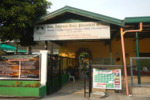 Der Eingang der San Lorenzo Ruiz Pfarrschule zeigt eine Tafel mit Text, einen Metallzaun, Menschen auf Stühlen unter einem Dach, Pfosten, ein Schild, Pflanzen, einen Baum und einen bewölkten Himmel.