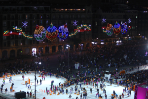 Eine große Menge von Menschen, die auf einer Eisbahn vor einem Gebäude Schlittschuhlaufen, beleuchtet von Straßenlaternen und geschmückt mit festlichen Weihnachtsdekorationen, mit Geländern und Pfosten, die die Bahn umgeben und einem dunklen Hintergrund.