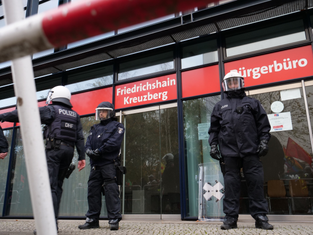 Polizeibeamte in Helmen und Handschuhen vor einem Glaswandgebäude mit angeklebten Papieren und einem Pfahl links, als "Friedrichshain Kreuzberg" Text auf Hintergrundtafeln erscheint.
