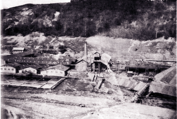 Schwarzes und wei├čes Foto eines Kohlebergbaus mit Geb├Ąuden, Masten und B├Ąumen im Hintergrund, darunter Text.