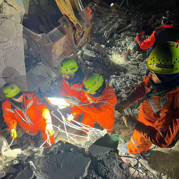 Rettungskräfte in orangefarbener Uniform und grüner Helm beseitigen Trümmern von einem eingestürzten Gebäude mit Werkzeugen, mit einem Kran im Hintergrund.