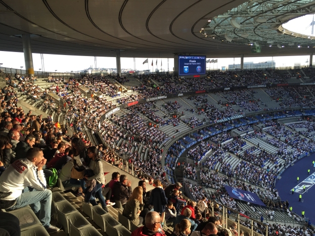 Große Menschenmenge in einem Stadion bei einem Fussballspiel, mit einer Bühne rechts, Fahnen, Stangen, einem Bildschirm und der Allianz Arena in München, Deutschland im Hintergrund.