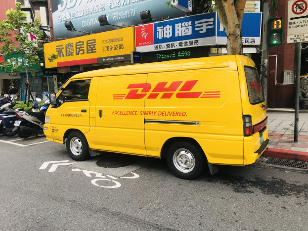 Gelbes DHL-Lieferfahrzeug auf der Straßenseite geparkt, umgeben von Gebäuden, Bäumen, Schildern, Mästen, Ampeln und einer Person auf dem Gehweg.