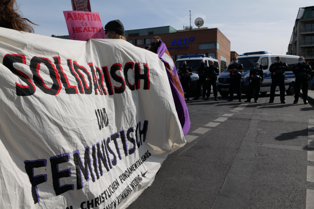 Eine Gruppe von Menschen marschiert auf einer Straße, hält ein "Solidarität und Feminismus"-Schild und hat parkende Fahrzeuge und Gebäude im Hintergrund unter einem klaren blauen Himmel.