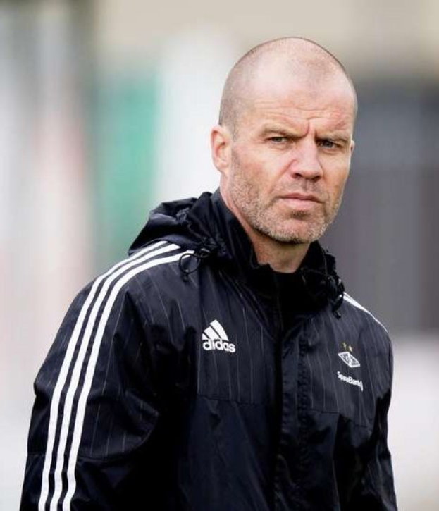 Bald Coach in einer marineblauen Jacke mit weißen Streifen und weißem Kragen, der auf einem Fußballfeld steht und die Hände verschränkt hält.