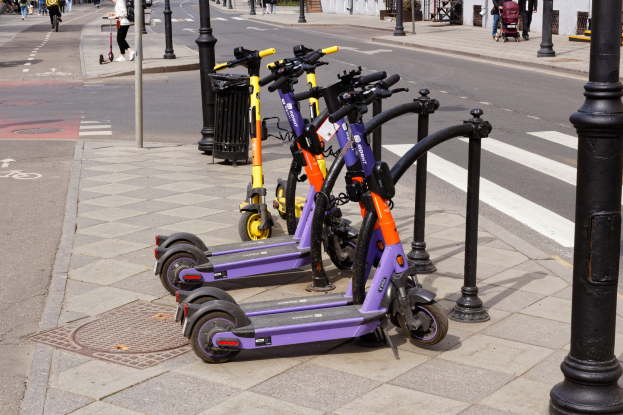Eine Reihe von Elektro-Scootern, die an der Seite einer Straße geparkt sind, mit Menschen, die auf dem Gehweg gehen und Fahrräder fahren, einem Müllcontainer im Vordergrund und Gebäuden, Bäumen und Pflanzen im Hintergrund.