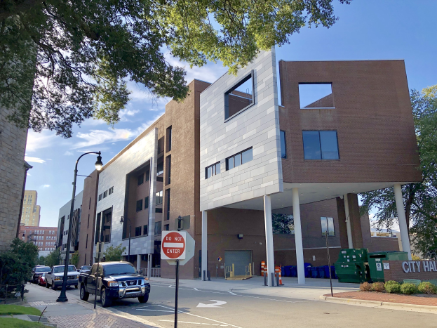Großes mehrstöckiges Rathausgebäude mit zahlreichen Fenstern, Straßeninfrastruktur einschließlich Masten, Laternen und Verkehrszeichen, umgeben von Fahrzeugen, Grünflächen und einem bewölkten Himmel.