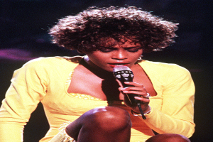 Whitney Houston performs bei den Grammys, trägt ein gelbes Kleid und hält ein Mikrofon in der Hand.
