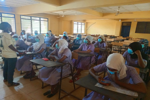 Ein Klassenzimmer in Nigeria mit Schülern, die Gesichtsmasken tragen und auf Bänken sitzen oder stehen, mit Büchern und Gegenständen auf Tischen, Fenstern, einer Tür, einer Wand, einer Decke mit Lampen und Ventilatoren und anderen Gegenständen im Hintergrund.