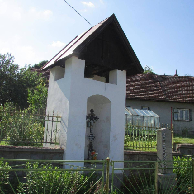 Kleines weißes Kirchengebäude mit einem Kreuz darauf, umgeben von einem Metallzaun, Pflanzen, Gras, einem Weg, nahen Häusern, Polen, Drähten und einem bewölkten Himmel, identifiziert als die Kirche des Heiligen Grabes.